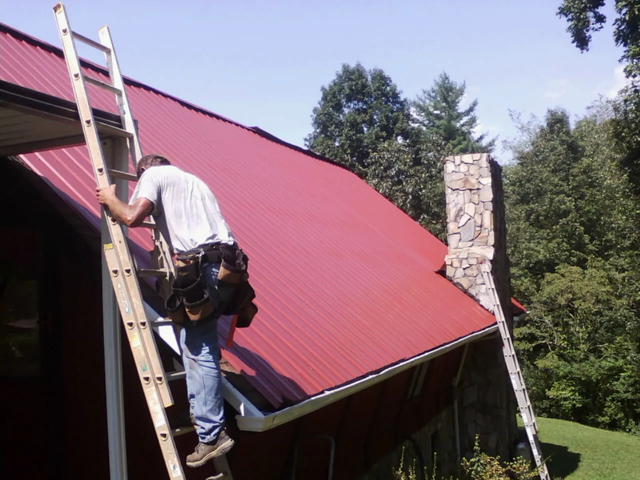 Roofer on a ladder beside a red metal roof and stone chimney — Soffit & Fascia Repair by Roofing Brothers in Glenarm